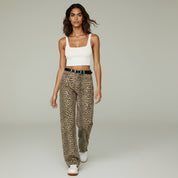 Woman wearing a white cropped tank top paired with high-waisted leopard print trousers and white sneakers, walking against a neutral studio background.