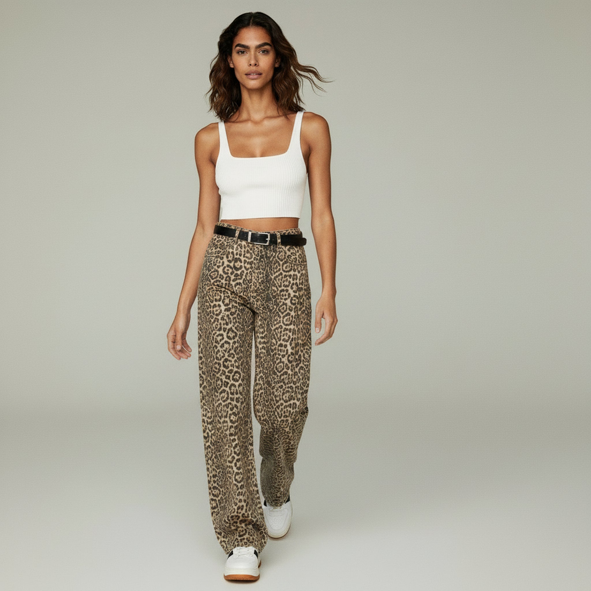 Woman wearing a white cropped tank top paired with high-waisted leopard print trousers and white sneakers, walking against a neutral studio background.