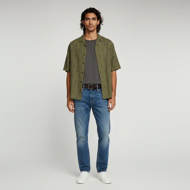 man wearing dark grey t-shirt with green polo shirt paired with soft blue jeans on a plain background