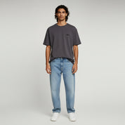 man wearing dark brown shirt and light blue jeans on a plain background